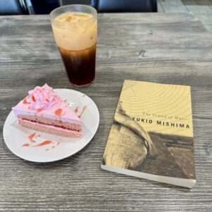 A photo of the book The Sound of Waves by Yukio Mishima with a slice of cake and a Thai iced tea
